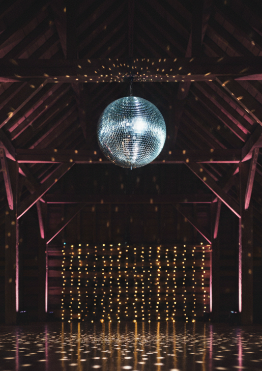 A polished wooden dance floor under a dramatic, mirror-tiled disco ball suspended from rustic barn rafters, with no dancers present. The disco ball scatters tiny reflections of light across the darkened space, while subtle, colored uplighting washes the timber beams in deep indigo and soft magenta. In the background, blurred fairy lights drape along the walls, adding warm golden bokeh. The camera is positioned low, angled slightly upward toward the disco ball, emphasizing its sparkle. The mood is glamorous and energetic, suggesting an unforgettable party, captured in crisp photographic realism with a cinematic, evening atmosphere.